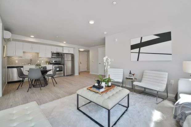Bright open-plan apartment interior combining a compact kitchen and dining area with a living room. The space features white flat-panel cabinetry, stainless-steel appliances, a pale tiled backsplash, light timber-look flooring, a tufted white ottoman, contemporary seating, and a large abstract black-and-white wall artwork.