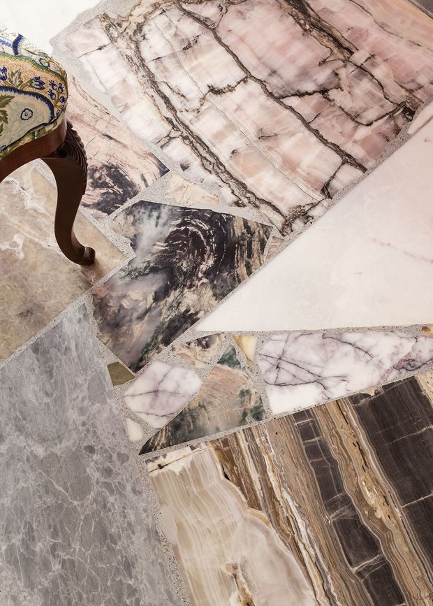 Close-up overhead view of a curated collection of large natural stone slabs or tiles with varied veining and mineral patterns, including pale pink, white, grey, black, beige, and brown tones. A carved wooden furniture leg with patterned upholstery appears at the upper left, providing scale and suggesting a showroom or interior materials display.