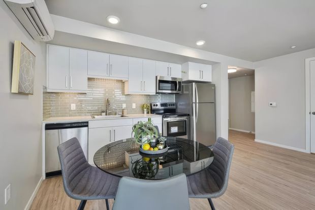 A bright, compact open-plan kitchen and dining area with white shaker-style cabinetry, stainless-steel appliances, a glossy beige horizontal tile backsplash, and light timber-look flooring. A round glass dining table with upholstered chairs occupies the foreground, while recessed ceiling lights and an overhead split-system air conditioner provide a clean contemporary fit-out.