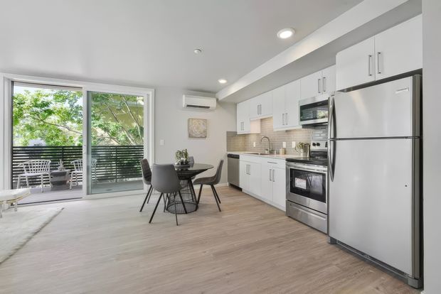 Bright open-plan apartment interior combining a compact white kitchen with a dining area and living space. The kitchen features white upper and lower cabinetry, stainless-steel appliances, a small tiled splashback, recessed ceiling lights, and pale wood-look flooring. Full-height glazed sliding doors open to a balcony with outdoor furniture and leafy views.
