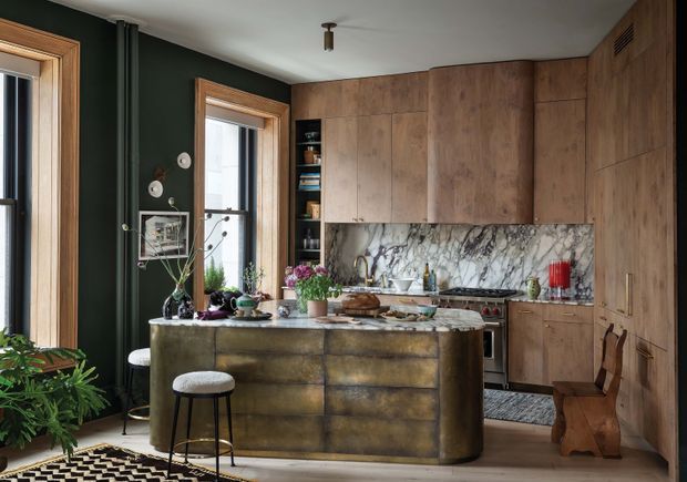 A richly styled kitchen with a large central island, dark green walls, warm timber-framed windows, extensive natural timber cabinetry, and a dramatic veined stone splashback. The island has a dark aged-metallic finish and includes casual seating, while open shelving, decorative objects, plants, and pendant-free track lighting add an eclectic, lived-in character. The space is compact to medium-sized with a standard-height ceiling and a warm, moody palette.