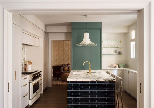 A refined kitchen viewed through a wide interior doorway, featuring white built-in cabinetry, a dark timber floor, a deep green central island with a white marble benchtop, and a dark navy-blue tiled island base. The island includes a brass tap and seating, while a sculptural pendant light hangs above. A concealed cooking area with a freestanding range is visible to the left, and open shelving with glassware lines the rear wall. The space has warm natural light and a traditional-meets-contemporary character.