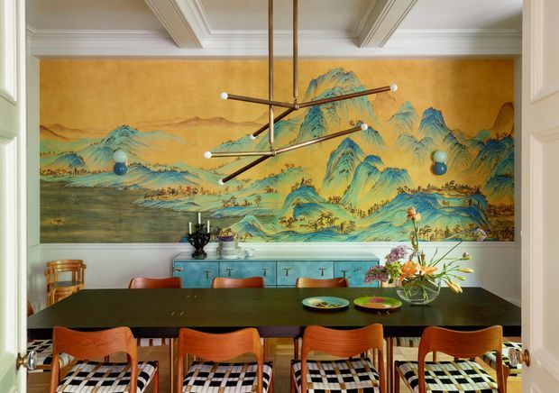 A richly styled formal dining room viewed through open double doors. A long dark dining table is surrounded by warm wood dining chairs with black-and-white checked upholstered seats. The rear wall features a large colourful chinoiserie-style landscape mural framed by pale green-grey wall panelling and white decorative ceiling mouldings. A turquoise built-in sideboard, sculptural brass-and-black linear pendant light, floral arrangement, and traditional accessories complete the eclectic, dramatic interior.