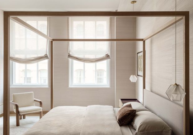 Bright, refined bedroom featuring a low-profile four-poster bed with dark timber framing, upholstered bedding, pale textured wall panels, tall windows with relaxed Roman shades, a light armchair, and a geometric pendant light. The room has a warm neutral palette and a clean, tailored contemporary character.