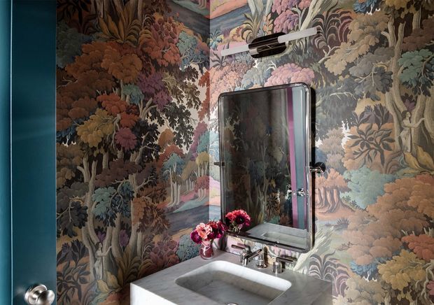 A compact bathroom vanity corner with a white rectangular basin, chrome tapware, framed mirror, and wall-mounted light. The walls are covered in richly patterned botanical forest wallpaper, contrasted by a deep teal painted door. The space has a dramatic, eclectic decorative character with soft, moody lighting.