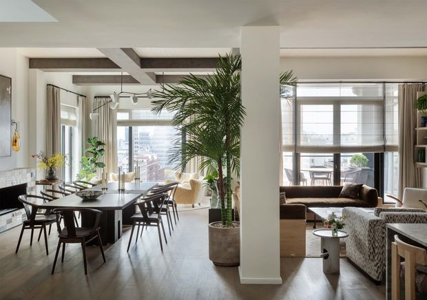 Open-plan apartment interior combining a dining area and living room, with floor-to-ceiling windows, city views, a large dark dining table, upholstered seating, abundant indoor palms, and a neutral contemporary palette. A substantial central column visually separates the dining and lounge zones, while exposed ceiling beams add architectural definition.
