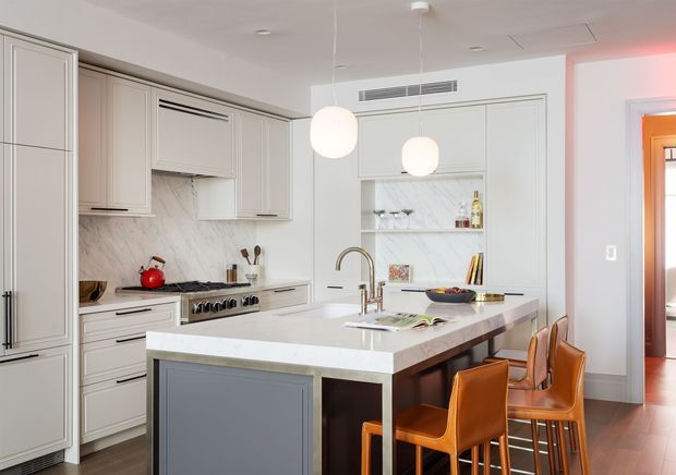Bright, refined kitchen with pale grey built-in cabinetry, a large marble-look island with integrated sink and brass-toned tapware, open shelving, and a contemporary range cooker. Warm caramel leather bar stools contrast with the light cabinetry, while pendant globe lights and timber flooring add warmth. A doorway at right provides a secondary view toward an adjoining room or hallway.