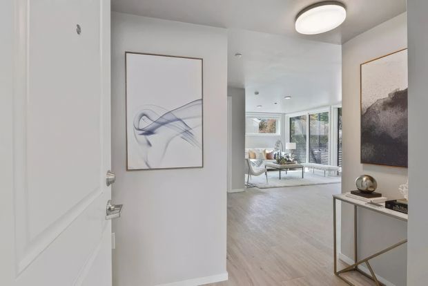 View from an entryway through an open white door into a bright, contemporary living room. The space features pale timber flooring, white walls and ceilings, recessed lighting, large glazed doors or windows, abstract wall art, and a slim console table with metallic legs and decorative objects.