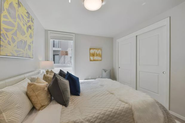Bright, compact bedroom with a neatly made bed, layered neutral and navy pillows, white quilted bedding, pale grey walls, white paneled double closet doors, a small window, bedside lamp, and understated abstract artwork. The room has a clean, calm contemporary styling with soft textures and restrained colours.