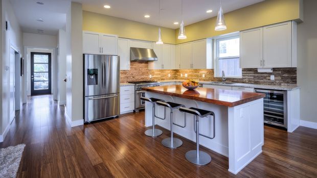 Bright contemporary kitchen with white shaker-style cabinetry, stainless-steel appliances, warm wood flooring, and a large central island topped with a richly stained timber benchtop. A rustic wood-look tiled or timber backsplash runs along the rear counters beneath recessed lighting and pendant lights. The island includes seating for four and opens to a hallway and exterior door.