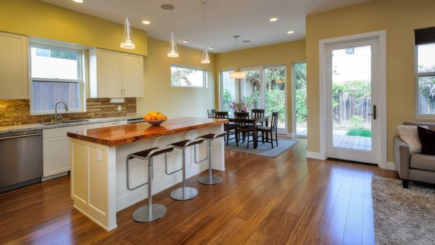 Open-plan kitchen and dining area with warm yellow walls, white cabinetry, a large timber-topped island with bar seating, patterned backsplash, pendant lighting, and dark timber flooring. Glazed doors and windows provide views to a small outdoor area, while a living area is partially visible at right.