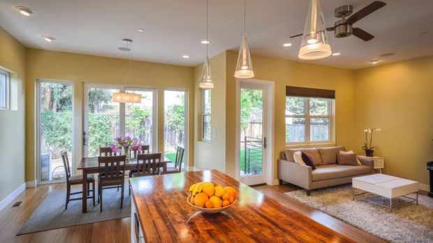 Open-plan living and dining area with warm yellow walls, polished timber flooring, a large glossy timber dining table, dark wood dining chairs, and a beige sofa on a textured rug. Tall glazed doors and windows overlook greenery, while pendant lights and a ceiling fan provide layered lighting and ventilation.
