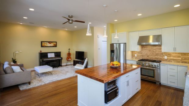 Open-plan contemporary kitchen and living area with a large timber-topped white island, stainless-steel appliances, pale cabinetry, and a warm stone or brick-pattern splashback. The adjoining living room includes a sofa, rug, piano, television, and ceiling fan, with timber flooring throughout and recessed lighting.