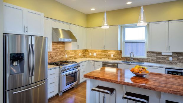 Bright contemporary kitchen with white raised-panel cabinetry, stainless steel appliances, a timber island benchtop with breakfast-bar seating, warm wood flooring, and a mixed stone and wood-look backsplash. Pendant lights and recessed ceiling lights illuminate the space.