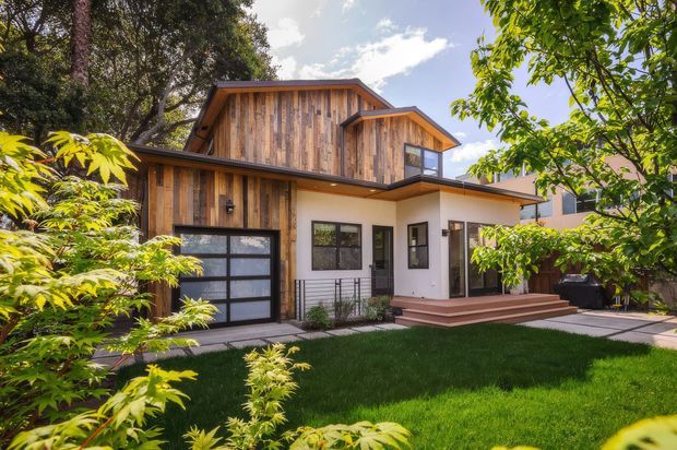 Exterior view of a contemporary two-storey house set within a leafy suburban garden. The facade combines warm vertical timber cladding on the upper level and garage wall with smooth white rendered walls, dark-framed glazing, a projecting flat roof, and a small timber entry deck and steps. Mature planting and lawn surround the home.