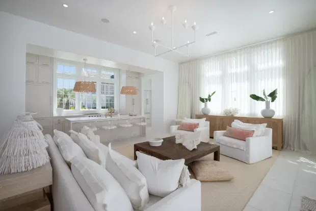 Bright, spacious open-plan living room with a refined coastal-inspired interior. The room features white upholstered sofas and armchairs, a dark timber coffee table, a pale textured rug, floor-to-ceiling sheer curtains, white built-in cabinetry around a large window, pendant lighting, and potted greenery. A glimpse of the adjacent kitchen or serving area is visible through the wide opening.