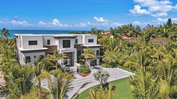 Aerial exterior view of a large modern white multi-storey residence set among dense tropical palms, with the ocean visible beyond. The home has a flat-roofed geometric form, extensive glazing, projecting upper-level volumes, paved driveway and landscaped grounds under bright blue skies.