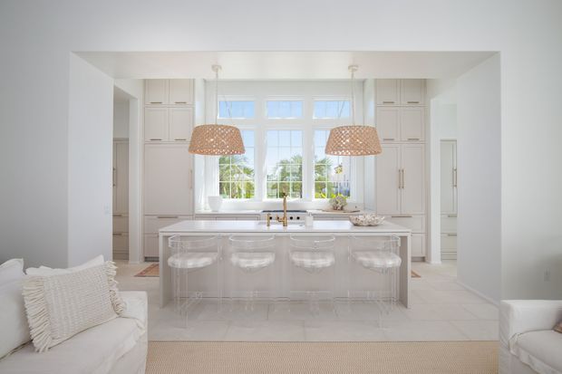 Bright, airy white kitchen with a large central island, integrated white cabinetry, pale tiled flooring, and a bank of multi-pane windows. The island has a light stone benchtop, transparent bar stools, and twin woven pendant lights. White upholstered seating is visible at the edges of the image, with a soft neutral rug in the foreground.