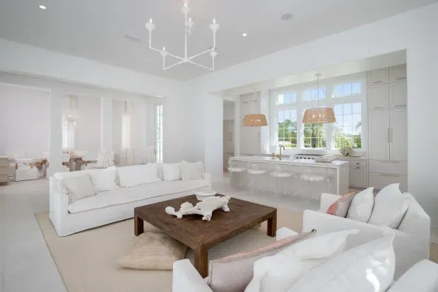 Bright, open-plan white living room flowing into a large kitchen. The room features white sofas, a substantial dark timber coffee table, pale neutral rugs and cushions, a white island with integrated seating, extensive light cabinetry, a large multi-pane window, woven pendant lights, and a contemporary linear chandelier. A dining area is visible beyond the living space.