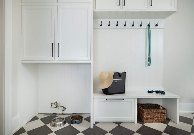Bright, compact mudroom or entry storage area with white built-in cabinetry, upper cupboards, a wall of black hooks, and a built-in bench with drawer storage. A black-and-white diamond-pattern tiled floor, woven basket, pet bowls, hat, and leash are visible. The room has white vertical wall panelling, crisp white trim, and a clean traditional-inspired fit-out.