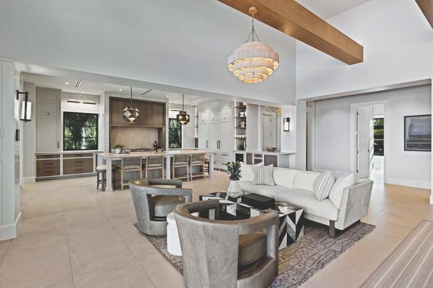 Bright open-plan interior combining a contemporary kitchen, dining island, and living room. The space features pale tiled flooring, white cabinetry and walls, a large light sectional sofa, sculptural lounge chairs, geometric rug, dark coffee table, pendant lighting, and a prominent exposed timber ceiling beam. Glazed doors and windows provide views to greenery outside.