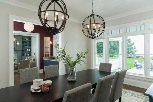 A refined dining room with a large dark timber dining table surrounded by upholstered grey dining chairs. Two oversized black metal spherical chandeliers hang from a textured white ceiling, while large white-framed windows provide views to a green outdoor setting. An open doorway reveals a dark blue-grey built-in bar or cabinetry area, and a deep burgundy feature wall adds contrast.