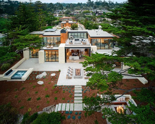 Elevated aerial view of a large contemporary residential home set within a landscaped suburban or bushland site. The house has a low, flat-roofed composition with extensive floor-to-ceiling glazing, pale masonry walls, warm timber-clad vertical elements, rooftop terraces, and multiple outdoor entertaining areas. Formal garden beds, stepping-stone paths, a small rectangular water feature, and an outdoor fire feature surround the building, while mature evergreen trees frame the property.