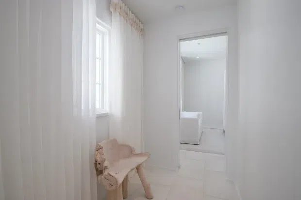 Bright, narrow white-painted interior passage or landing with vertical wall panelling, a tall window dressed in sheer curtains, pale tiled flooring, and a small pale upholstered bench. An open doorway leads to another bright room with a partially visible white bathtub or fixture. The space has a clean, airy, minimalist-traditional feel.