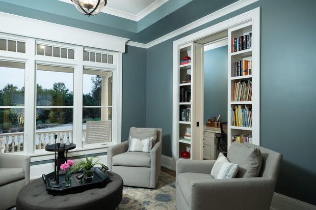 A refined sitting room with deep blue-grey walls, crisp white architectural trim, large traditional multi-pane windows, upholstered armchairs, a round ottoman coffee table, and built-in bookcases flanking a doorway to a small study or workspace. The room has a calm, classic character with views to a landscaped exterior.