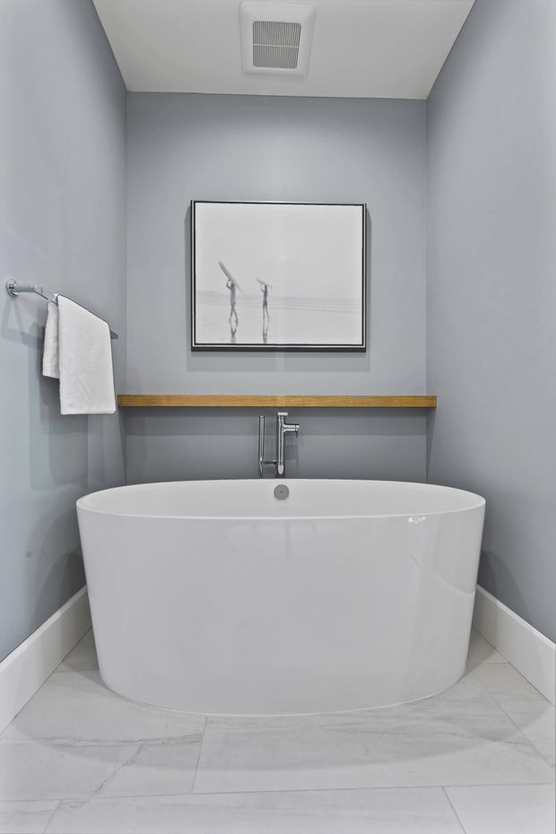 A compact, refined bathroom featuring a freestanding white oval bathtub beneath a wall-mounted chrome faucet. The room has cool light-grey painted walls, pale stone-look floor tiles, white skirting, a slim timber shelf, a framed monochrome artwork, and a towel rail with a white towel. A ceiling ventilation grille is visible above.
