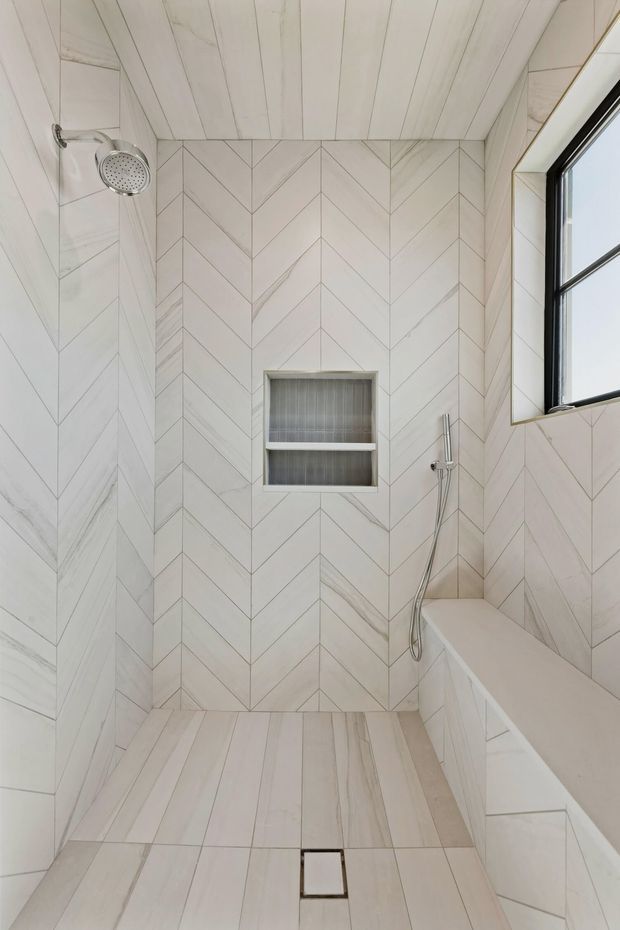 Bright, compact walk-in shower with pale marble-look tiles arranged in a chevron pattern on the walls and bench surround, and linear plank-style tiles on the ceiling and floor. The shower includes a recessed tiled niche with two shelves, a wall-mounted showerhead, handheld shower, built-in bench, dark-framed window, and square floor drain. The overall design is clean, refined, and highly cohesive in light neutral tones.