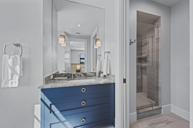Bright contemporary bathroom with a navy-blue vanity, marble-look countertop and integrated basin, large wall mirror, twin wall sconces, towel ring, and a glass-enclosed tiled shower. A doorway and reflected bedroom or hallway space are visible beyond the vanity.