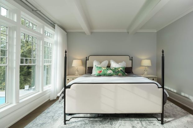 Bright, refined bedroom with a black metal four-poster-style bed, white bedding, bedside tables and lamps, and large multi-pane windows along one wall. The room features pale grey walls, white coffered ceiling beams and trim, dark timber flooring, and a light patterned area rug. Garden greenery and a glimpse of a pool are visible outside the windows.