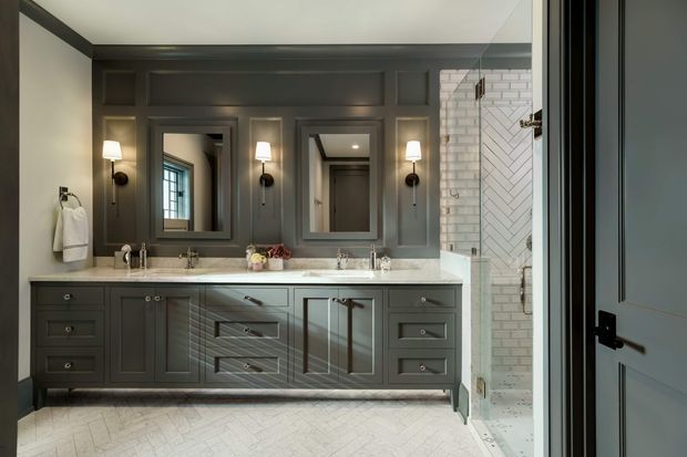 A refined double-vanity bathroom with dark grey-blue painted panelled cabinetry, marble-look countertop, two framed mirrors, wall sconces, and a white subway-tiled shower enclosure with herringbone detailing. The light herringbone floor and bright natural light create a polished, traditional interior.