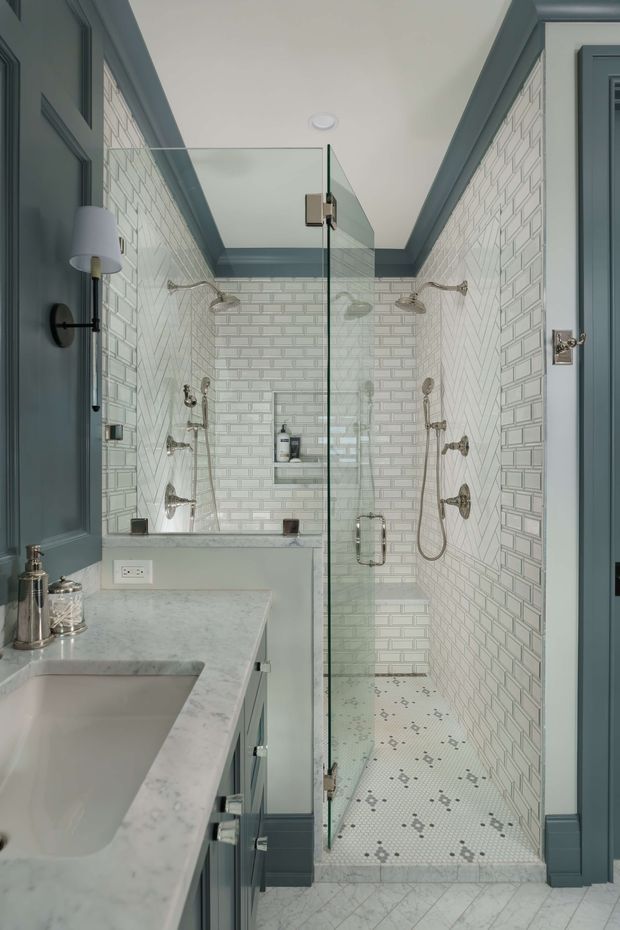 A narrow, traditional-style bathroom featuring a walk-in glass shower enclosure with dual shower fixtures, white subway-tile walls, and a small white-and-grey mosaic shower floor. The room has dark blue-grey painted timber-panelled cabinetry and trim, a pale marble vanity benchtop with undermount basin, brushed metal hardware, wall sconces, and a light patterned floor tile.