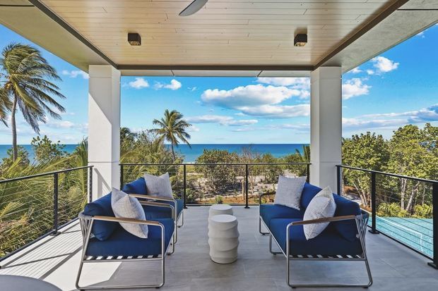 A covered elevated outdoor lounge terrace overlooking a tropical coastline and ocean. Two navy lounge chairs with pale cushions face each other around a small sculptural white table. The terrace has grey tiled flooring, slender dark metal balustrades, white structural columns, and a timber-lined ceiling with recessed lighting and a ceiling fan. Palm trees, turquoise water, and bright sky form the background.