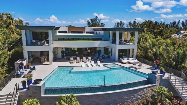 A large contemporary two-storey residence set amid lush tropical landscaping, viewed from the pool side. The building has crisp white rendered walls, extensive glazing, projecting flat roof planes, deep shaded balconies, and a broad elevated terrace. A geometric turquoise swimming pool with an integrated spa dominates the foreground, surrounded by pale stone or concrete paving, loungers, and planted areas.