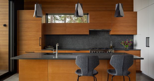 Contemporary kitchen with warm timber cabinetry and wall panelling, a dark stone-look splashback and island benchtop, integrated appliances, white full-height storage, and two upholstered bar stools. Three geometric pendant lights hang above the island, while a narrow window brings in views of trees.