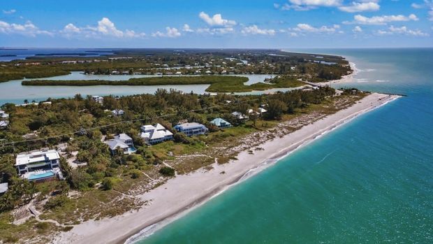 Aerial coastal view of a narrow beachfront settlement, with turquoise ocean, a sandy shoreline, scattered houses, waterways and lush vegetation across a low-lying coastal landscape.