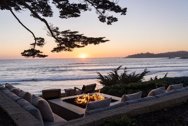 A coastal outdoor lounge terrace overlooking the ocean at sunset, furnished with cushioned built-in seating arranged around a square fire pit. Dark timber decking or paving and low coastal landscaping frame the view, with silhouetted windswept trees overhead and a headland across the water.