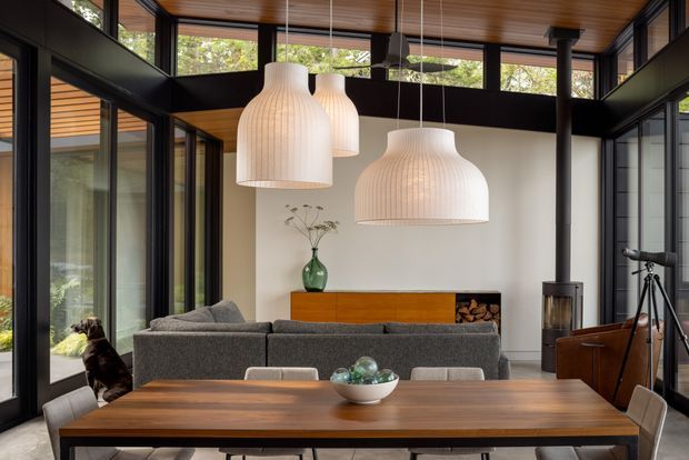 A warm, contemporary open-plan living and dining space with extensive black-framed glazing, a timber-lined ceiling, and a large timber dining table in the foreground. A grey sectional sofa faces a simple fireplace wall with timber joinery and stacked firewood, while oversized ribbed pendant lights hang above the dining area. The room has a strong indoor-outdoor connection and a refined, natural material palette.