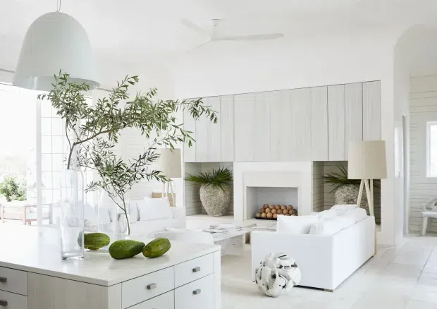 Bright, airy white living room with a large built-in timber-toned wall storage installation surrounding a recessed fireplace. White sofas, pale flooring, a central white island or console with drawers, sculptural pendant lighting, floor lamps, and natural greenery create a clean contemporary interior. An adjacent bright room is visible through the opening at right.