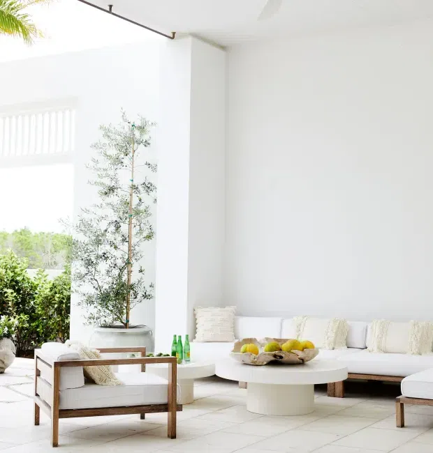 Bright, airy white living room with a minimalist coastal-inspired interior. The space features white built-in or low-profile seating with textured neutral cushions, a rounded white coffee table, timber-framed lounge chairs, pale tiled flooring, large open glazing, and indoor greenery. The high white walls and abundant daylight create a serene, spacious atmosphere.