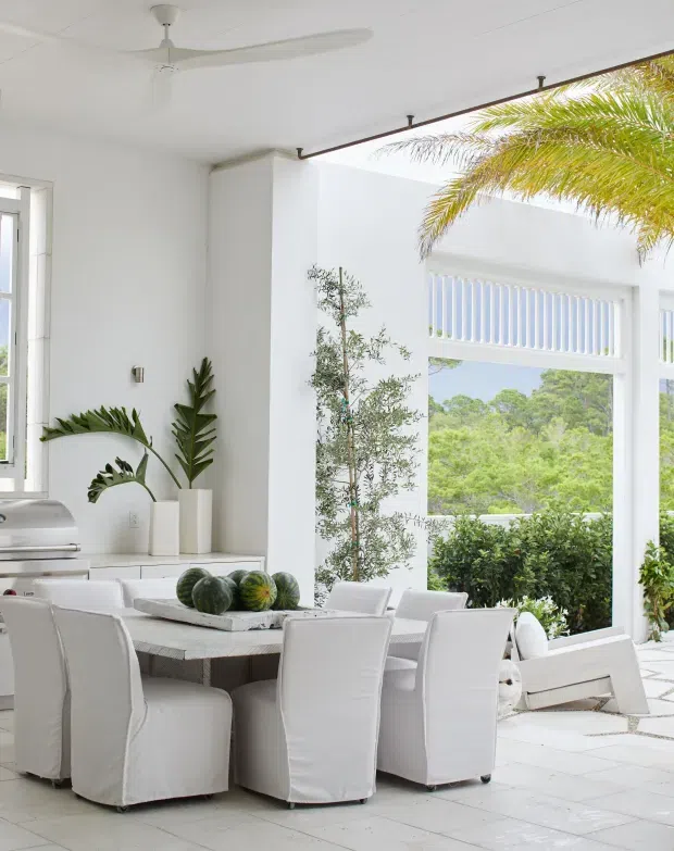 Bright, airy dining area with a large white dining table surrounded by slipcovered white chairs. The predominantly white interior has large windows and an open glazed wall overlooking lush greenery, white tiled flooring, a ceiling fan, potted tropical plants, and a partial outdoor patio visible beyond. A barbecue or outdoor cooking appliance is visible at the left edge.