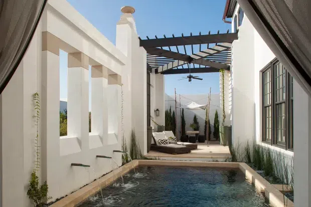 A narrow, sunlit courtyard pool framed by white rendered walls and tall architectural openings. A dark timber pergola with a ceiling fan shades an outdoor lounge at the far end, while a small rectangular pool, pale stone coping, potted plants, and climbing greenery create a private resort-like setting. Mountain scenery is visible beyond the courtyard.