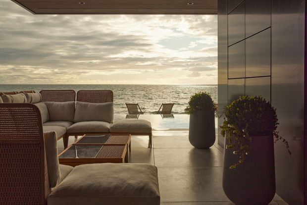 A sophisticated indoor-outdoor lounge opening directly onto a sandy beach and ocean. The space features a neutral upholstered sectional and woven lounge furniture, a low timber-and-glass coffee table, large dark planters, smooth stone or concrete flooring, dark built-in wall panelling, and a timber-lined ceiling. The open glazing frames a calm coastal view with additional outdoor seating near the water.