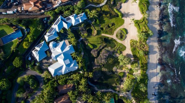 Aerial view of a large coastal resort or residential compound with multiple interconnected buildings featuring bright white pitched and faceted roofs, set within lush tropical landscaping beside a sandy beach and rocky ocean shoreline. Curving pathways, lawns, palm trees and landscaped garden areas surround the buildings.