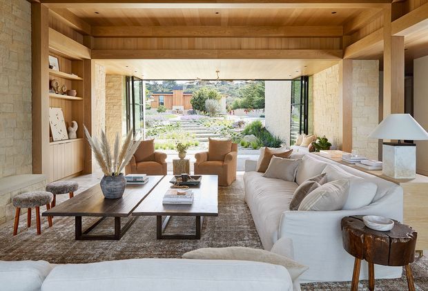 A warm, refined living room framed by exposed timber beams and pale textured stone walls. The room features a large cream sectional sofa, sculptural armchairs, a timber-and-metal coffee table, built-in timber shelving, and broad glazed openings overlooking a landscaped garden. Natural materials, abundant daylight, and the strong indoor-outdoor connection create a relaxed contemporary setting.