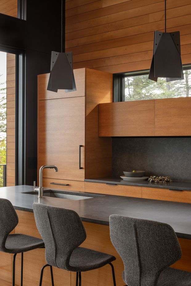 A sophisticated modern kitchen featuring extensive warm timber wall panelling and cabinetry, a dark stone or composite benchtop and splashback, black-framed glazing, and sculptural dark pendant lights. The island includes an undermount sink with a brushed metal tap and seating for several people on upholstered charcoal bar stools. Natural light enters through large windows, creating a warm, refined atmosphere.