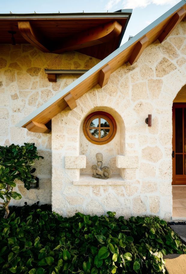 Close-up exterior view of a stone-clad building featuring intersecting pitched rooflines, exposed timber eaves and gable trim, a round timber-framed window, and a recessed decorative wall niche. Dense green planting borders the base of the facade.